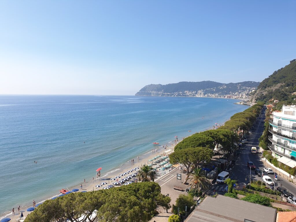 Sunny coastal scene with a sandy beach, turquoise sea, and hillside town along the shore.