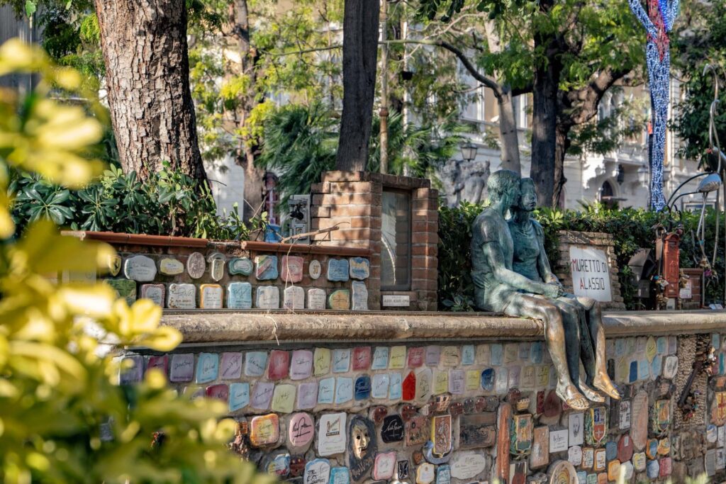 Colorful tiled wall at Muretto di Alassio with a bronze seated sculpture and trees in a garden setting.