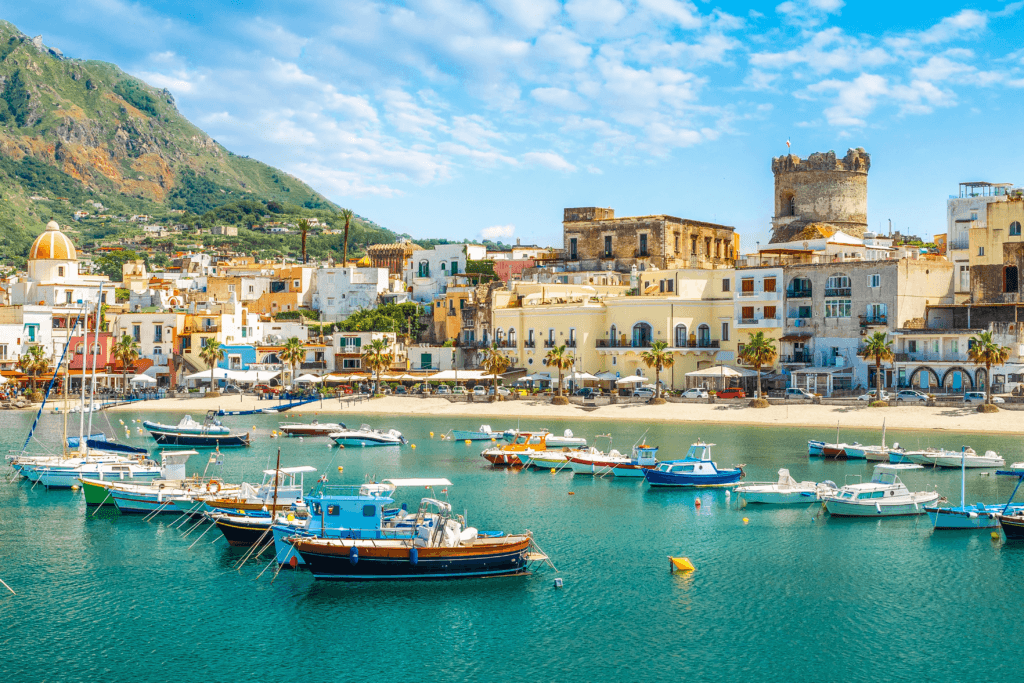 Harbor view of a Mediterranean town with colorful buildings and boats anchored in calm turquoise water.