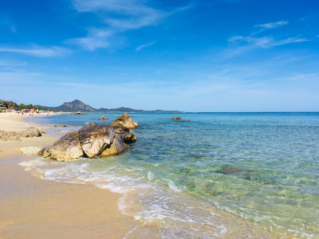 Sunny tropical beach with turquoise water, rocky outcrop and people lounging along the shore under a bright blue sky.