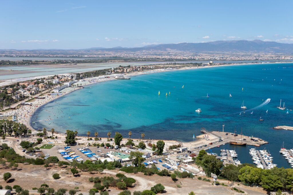 Aerial view of a sunny seaside town with turquoise bay, sandy beach, boats, and distant mountains with blue sky.