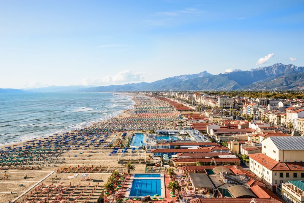 Crowded beach with colorful umbrellas and sunbathers along the shoreline, mountains in the distance behind a seaside town.