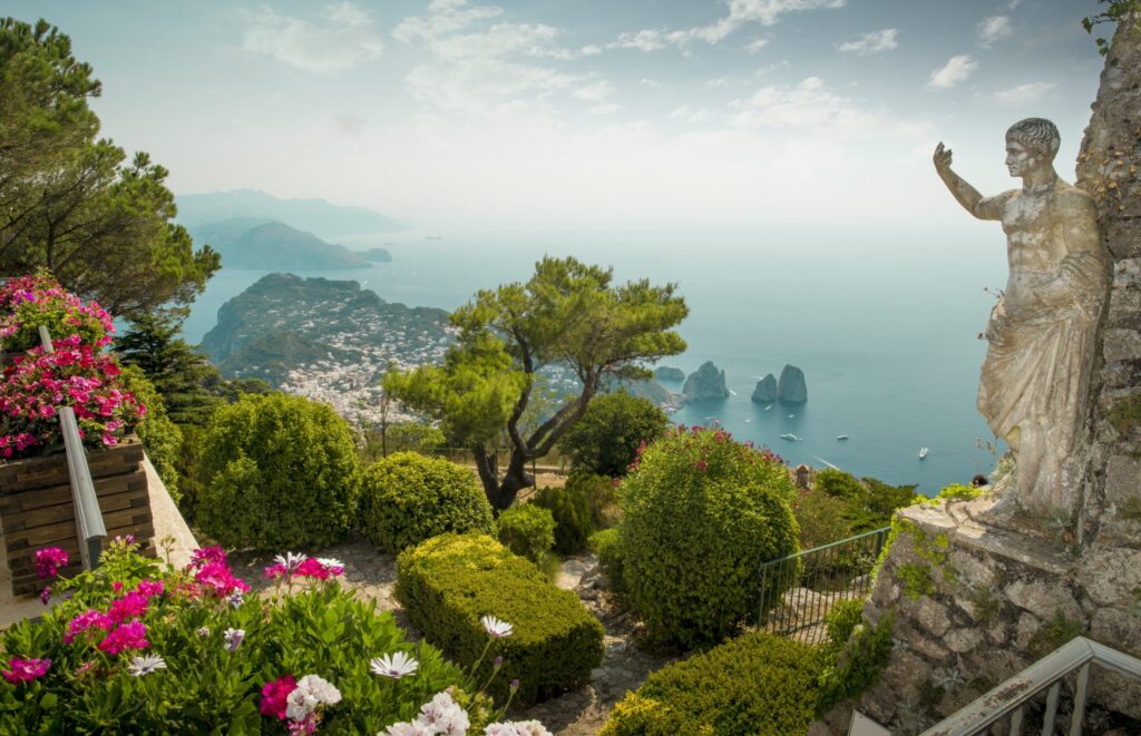 Garden foreground with pink and white flowers, overlooking a coastal town and blue sea to the left; a stone statue stands on the right.